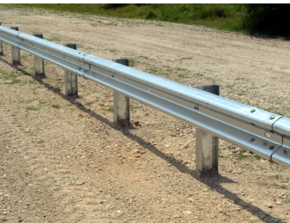 Galvanized steel W-Beam highway guardrail installed on driven posts along a roadside dirt shoulder, showing the corrugated rail profile and splice connections.