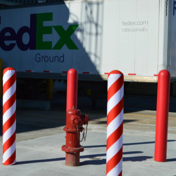  Red and white spiral-striped bollard covers installed at a FedEx Ground loading dock, protecting a fire hydrant and surrounding area from vehicle contact. 