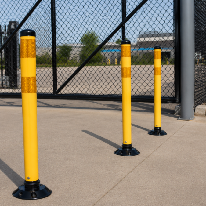 Yellow delineator posts with reflective bands installed on concrete in front of a chain-link security gate. 